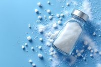 Glass jar filled with white salt or sugar crystals scattered on blue background, minimalist food photography composition with soft shadows and light.
