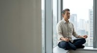 Middle-aged Asian man meditating in lotus pose on office windowsill with city skyline view, finding mindfulness and calm during work break.