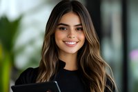 Young Hispanic woman with long wavy brown hair and bright smile wearing black top, holding smartphone against blurred background. Professional headshot for business.