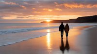 Romantic couple silhouettes walking hand in hand on golden sunset beach with dramatic orange sky clouds reflecting on wet sand and gentle ocean waves.