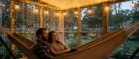 Couple relaxing in hammock under warm string lights in cozy screened porch at dusk, enjoying evening together in comfortable outdoor retreat space.