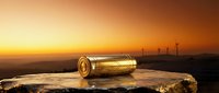 Metallic gold cylinder rests on rocky surface at sunset with wind turbines silhouetted on horizon, symbolizing renewable energy and sustainability concepts for environmental campaigns.