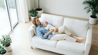 Young blonde woman relaxing on white sofa with smartphone in bright modern living room with green plants and natural light from large windows.