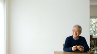 Senior man with gray hair and glasses, wearing dark blue sweater, sitting at wooden table with hands gesturing while speaking. Bright interior with white walls and natural light. Suitable for business, education, communication, and lifestyle content.