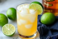 Refreshing whiskey sour cocktail with lime garnish and ice cubes on marble surface with fresh limes and bottle in background for bar menu designs.