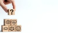Hand placing wooden cube with question mark above pyramid of blocks featuring wellness symbols including brain, heart, lotus, scales and smiley face.