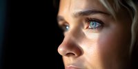 Close up profile portrait of young Caucasian woman with bright blue eyes and natural makeup looking away against dark background, dramatic side lighting.