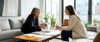 Two women in business casual attire discuss documents at a wooden table in a bright, modern office with plants and large windows, suitable for corporate, teamwork, and professional communication concepts.