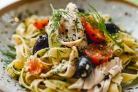 Fresh fettuccine pasta with shredded chicken, cherry tomatoes, black olives and dill garnish in creamy sauce on ceramic plate, macro food photography.