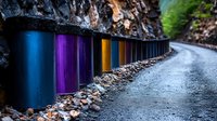 Colorful safety barriers line curved mountain road with vibrant blue, purple and orange posts against rocky cliff wall. Wet asphalt creates dramatic perspective.