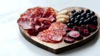 Heart-shaped wooden board with cured meats, salami, olives and nuts for romantic dinner or Valentine's Day charcuterie platter on white background.