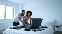 Young couple packing luggage on bed in bright bedroom. Man with beard and woman with curly hair organizing travel items for vacation or relocation.