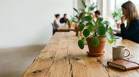 Young woman with brown hair and light skin tone, wearing dark clothing, works at wooden desk with potted plant in focus, notebook, and mug in bright office space with colleagues and greenery.