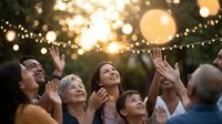 Multigenerational family celebrating outdoors at sunset, laughing and reaching up toward glowing string lights in a garden setting.