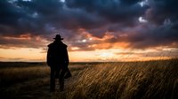 Silhouette of man in hat standing in golden wheat field at dramatic sunset with stormy clouds overhead creating moody atmospheric landscape scene.