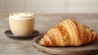 Fresh baked flaky croissant on wooden serving board with cappuccino cup in background, morning breakfast setting against blurred rustic surface.