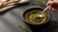 Ground green herbs and spices in dark bowl with spoon on rustic wooden table. Fresh seasoning powder for cooking and culinary preparations.