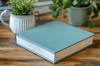 Mint green hardcover book on wooden table with potted plants in background, creating cozy home office or reading nook atmosphere. Minimalist composition.