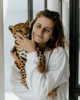 Young Caucasian woman in white sweater holding baby leopard cub near window in natural light, creating tender moment between human and wild animal. Gentle expression and soft mood.