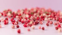 Pink and red dried peppercorns scattered on pink background, selective focus on aromatic spices creating minimalist culinary composition.
