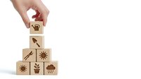 Hand placing wooden cube with watering can icon on pyramid of blocks featuring gardening symbols isolated on white background for design projects.