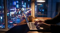 Professional working late on laptop computer in modern office with city lights view through window at night.