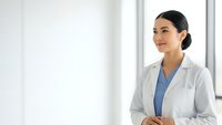 Professional female doctor in white coat smiling confidently in modern medical office with natural lighting for healthcare marketing materials.