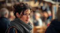 Young woman with curly hair and gray scarf looking thoughtfully in crowded marketplace, soft bokeh background creates atmospheric urban scene.