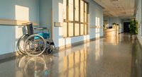 Empty wheelchair in modern hospital corridor with sunlight streaming through windows creating warm reflections on polished floor.
