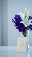 Elegant birthday flower arrangement with purple orchids and white lilies in glass vase beside greeting card on marble surface for celebration decor.