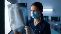 Female doctor in medical mask examining chest x-ray film in hospital. Healthcare professional analyzing lung radiograph for diagnosis and treatment planning.