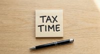 Yellow sticky note with tax time reminder message and black pen on wooden desk surface for financial planning and accounting preparation.