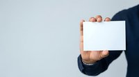 Businessman in dark navy suit holding blank white business card or paper against neutral gray background, cropped view of hand and arm for corporate branding and identity mockup.