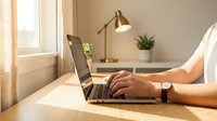 Professional workspace with person typing on laptop computer at wooden desk with modern brass lamp and plant creating productive home office environment.