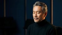 Mature Asian businessman with gray hair in black turtleneck looking down thoughtfully in modern office setting with warm lighting for corporate content.