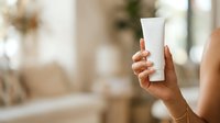 Woman with medium skin tone holding blank white cosmetic tube for skincare, lotion, or sunscreen product mockup in bright home interior setting.