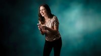 Young woman with long brown hair laughing while using smartphone against teal gradient background. Casual portrait for social media and technology concepts.