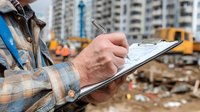 Construction worker writing on clipboard at building site with residential towers and equipment in background for project management and documentation.