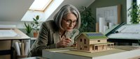 Mature woman architect wearing glasses examining wooden house model on desk in bright studio office space with plants and blueprints.