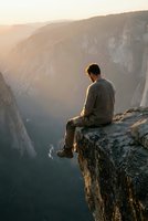 Young man sitting on cliff edge overlooking mountain valley during golden hour sunset, contemplating nature's beauty in peaceful solitude.