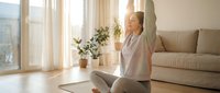 Woman in casual athletic wear practicing yoga stretching exercises on mat in bright modern living room with natural sunlight, potted plants, and comfortable furniture for wellness and home fitness.