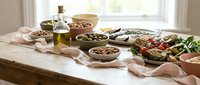 Mediterranean feast with olives, olive oil, fresh salad and rustic bowls on wooden table near bright window for healthy cooking and dining concepts.