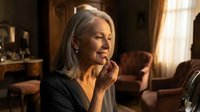 Mature blonde woman smiling warmly while talking on phone in cozy living room with soft natural lighting creating intimate domestic atmosphere.
