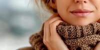 Close-up of Caucasian woman's face with natural makeup wearing cozy knitted brown scarf in winter setting, soft blurred background.