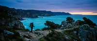 Hiker standing on rocky cliff overlooking turquoise Mediterranean cove at twilight, scenic coastal landscape for travel and adventure themes.