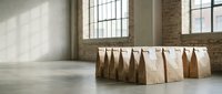 Row of brown paper lunch bags arranged on concrete floor in bright industrial loft space with brick walls and large windows for food delivery concept.