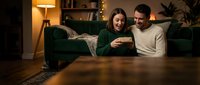 Happy young couple relaxing together on couch at home in evening, looking at smartphone and smiling in cozy living room with warm lighting for lifestyle content.