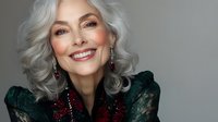 Elegant mature woman with silver gray hair wearing emerald green lace blouse and burgundy accessories smiling confidently at camera against dark background.
