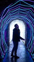 Young man walking through futuristic neon light tunnel with colorful LED strips creating geometric patterns in urban nighttime setting for technology concepts.