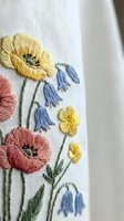 Delicate floral embroidery on white linen fabric featuring colorful wildflowers including poppies and bluebells with intricate stitching details.